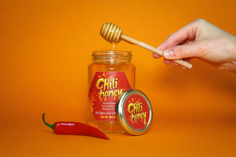 honeystickers-Event-Spicy-Honey Jar of Chilli honey with a wooden dipper, chili pepper, and honeycomb on an orange background