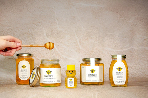 honeystickers_vaeriety-of-Charming-charly_design Honey jars and a bear-shaped honey container on a wooden surface with a neutral background.
