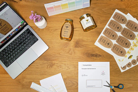 honeystickers_Header_creativity Laptop on a wooden desk with honey jars, labels, and stationery items.
