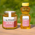honeystickers_Soft-Sophie-Hexagonal-Bear-Jar-Sustainability Jar and bear-shaped bottle of Blossom Honey on a wooden surface with a blurred green background